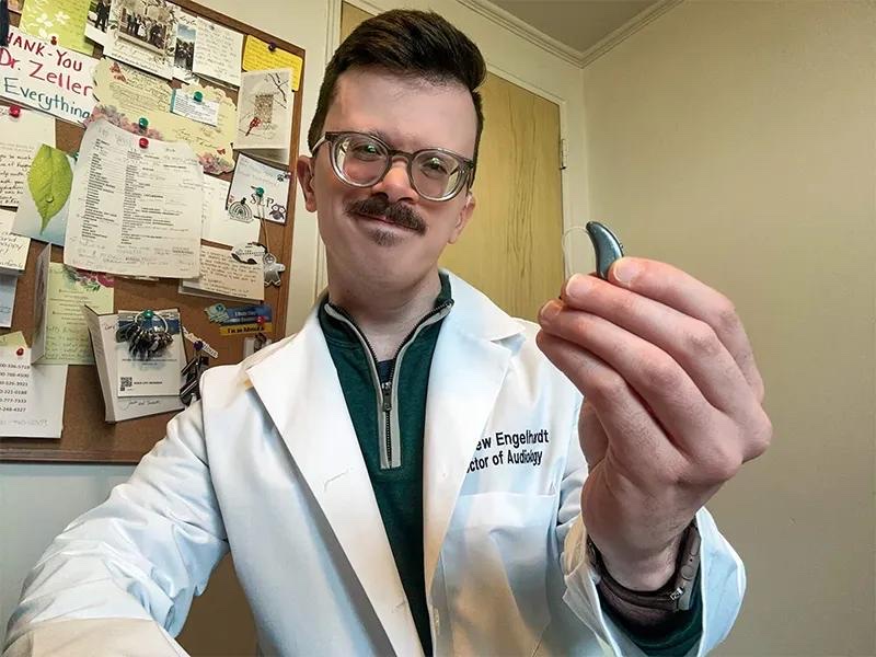 Dr. Engelhardt in his white coat holding up a hearing aid in his office